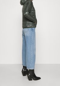 Zelená péřová bunda s kapucí, doplněná o světle modré džínové rovné kalhoty a černé kotníkové boty na podpatku. Hladká textura s lesklým povrchem.