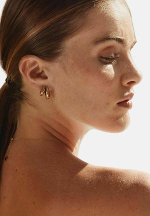 Jeune femme aux cheveux bruns attachés, portant des boucles d'oreilles en anneaux torsadés en or et un collier en or délicat, de profil contre une lumière vive.