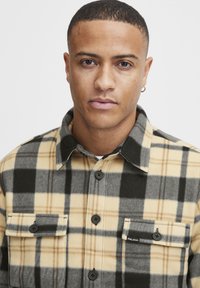 Checkered flannel shirt featuring black, gray, and beige colors, with long sleeves, buttoned chest pockets, and a collar.