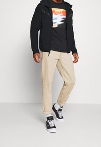 Man i ljusbeige uppvikta byxor, svart Nike T-shirt med grafik, svart zip-up hoodie, vita strumpor och svarta och vita Nike sneakers.