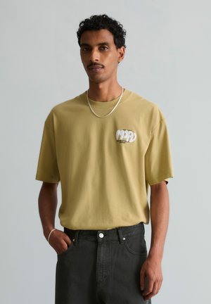 Mann mit lockigem Haar, der ein senfgelbes T-Shirt mit Logo und schwarze Hose trägt, steht vor einem schlichten hellen Hintergrund.