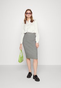 Witte blouse met lange mouwen, grijze geribbelde kokerrok, zwarte chunky loafers, met een kleine groene handtas en rode en grijze oversized zonnebril.