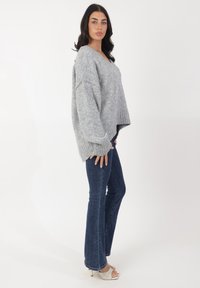 Grauer Strickpullover mit tiefem V-Ausschnitt, oversized Passform und geripptem Saum; kombiniert mit dunkelblauen Schlagjeans und weißen Absatzsandalen.