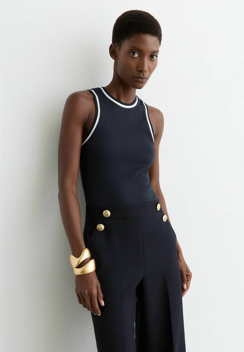 Vrouw met kort haar, gekleed in een mouwloze zwarte top met witte bies, zwarte broek met gouden knopen en een dikke gouden armband, staand tegen een witte muur.