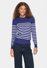 Pull rayé bleu marine avec des rayures blanches horizontales, col rond, et poignets et ourlet côtelés. Associé à un pantalon sombre.