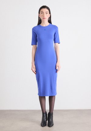 Modèle portant une robe bleue ajustée à manches courtes et col rond, complétée par des bottines noires et des boucles d'oreilles en créoles, se tient devant un fond uni.