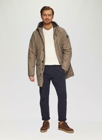 Beiger Parka mit Kapuze, schwarzer Fütterung, zwei Vorderentaschen; kombiniert mit einem cremefarbenen Pullover, navyfarbenen Hosen und braunen Turnschuhen.