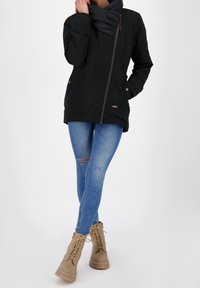 Czarna kurtka na zamek z szalikiem w kolorze szarym, niebieskie podarte obcisłe jeansy i beżowe sznurowane buty z brązowymi akcentami i nowoczesnym wzornictwem.