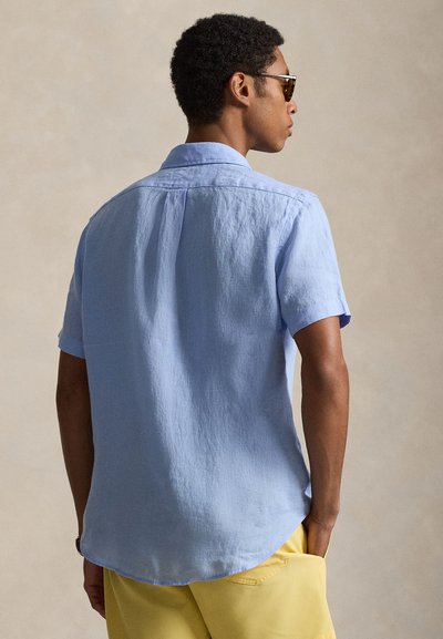 Άνδρας που φοράει ανοιχτό μπλε κοντομάνικο πουκάμισο και κίτρινα σορτς, εμφανίζεται από πίσω, με το ένα χέρι στην τσέπη και φορώντας γυαλιά ηλίου.