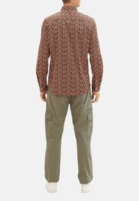 Gestreepte lange mouw shirt in bruin met veelkleurige ontwerpen; gecombineerd met lichtgroene cargobroek. Witte schoenen maken de outfit af.