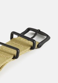 Bež pleteni remen s tri crne plastične kopče. Teksturirani materijal s detaljima šivanja. Jednostavan, podesivi dizajn.