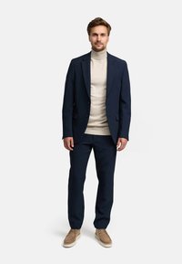 Abito blu navy con giacca e pantaloni su misura, abbinato a un dolcevita beige. Le scarpe sono in suede beige con una suola bianca spessa.