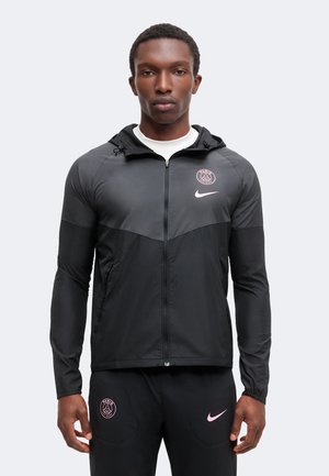 Homme portant une veste de sport noire Nike Paris Saint-Germain et un pantalon avec des logos roses, debout devant un fond clair uni.