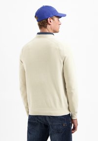 Beige gebreide trui met een hoge kraag, geribde details op de schouders en lange mouwen. Gecombineerd met donkerblauwe spijkerbroek en een blauwe pet.