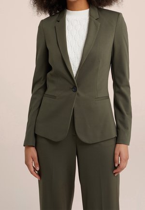 Femme portant un blazer vert olive ajusté avec un seul bouton noir, associé à un pantalon assorti et un haut en maille blanche texturée.