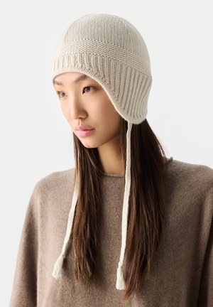 Junge Frau mit langen dunklen Haaren, die eine beige gestrickte Mütze mit Ohrenschützern und einen braunen Pullover trägt, leicht nach unten und zur Seite blickend.