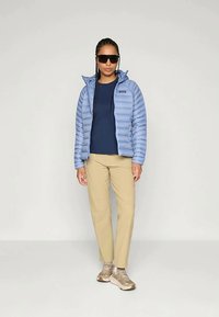 Žena u svijetloplavoj puf jakni, tamnoplavoj majici, bež hlačama, svijetlosmeškim tenisicama i velikim crnim naočalama stoji pred jednostavnom svijetlom pozadinom.