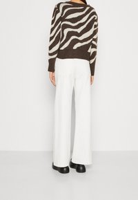 Woman wearing a brown and beige zebra-striped sweater, white wide-leg pants, and black shoes, standing facing a white wall.