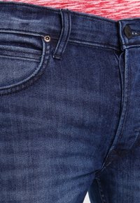 Mörkblå denimjeans med en slät yta, en klassisk femfickdesign och en knappstängning i midjan. Detaljer inkluderar sömnadsdekorationer.