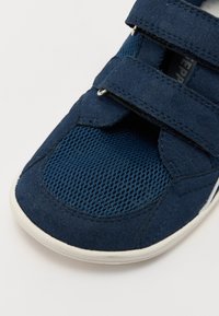 Sapatilha azul com fechos em velcro, uma combinação de camurça e materiais de malha, bico redondo e sola branca em borracha.