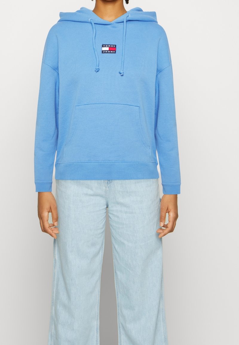 Personne portant un sweat à capuche bleu clair avec poche avant et écusson logo, associé à un jean large délavé clair.