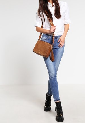 Bruine faux suede handtas met franjeretail, rechthoekige vorm, verstelbare schouderriem, gecombineerd met blauwe skinny jeans en zwarte veterschoenen.