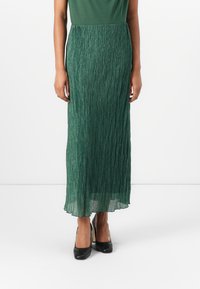 Gonna maxi plissettata verde con una finitura metallica, dotata di una vita liscia e una silhouette fluida, abbinata a scarpe con tacco alto nere.