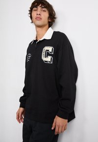 Sort polo skjorte med lange ærmer og hvid krave, som har et stort geometrisk bogstav "C" og mønstrede detaljer på forsiden.