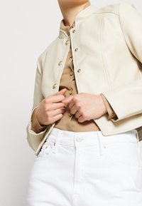 Veste en similicuir couleur crème avec col mandarin et boutons dorés, associée à une chemise beige et un pantalon en jean blanc.
