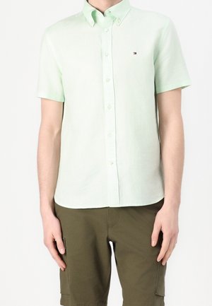 Uomo che indossa una camicia verde chiaro a maniche corte con bottoni e pantaloncini cargo verde oliva, in piedi con le braccia rilassate.