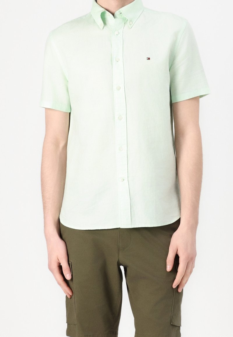 Homme portant une chemise vert clair à manches courtes boutonnée et un short cargo vert olive, debout les bras détendus.