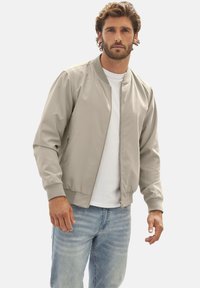 Hellgraue Bomberjacke mit Reißverschluss, gerippten Bündchen und Kragen. Verfügt über zwei Seitentaschen, getragen über einem weißen T-Shirt und blauen Jeans.