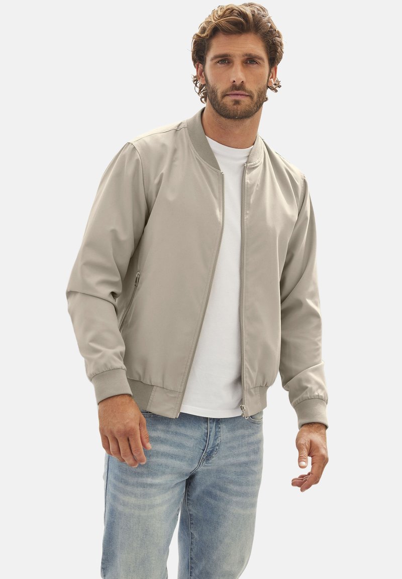 Hellgraue Bomberjacke mit Reißverschluss, gerippten Bündchen und Kragen. Verfügt über zwei Seitentaschen, getragen über einem weißen T-Shirt und blauen Jeans.