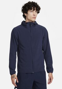 Marinblå lättviktsjacka med luva, dragkedja, justerbara dragsnören och sidofickor. Slät yta med en diskret logodetalj.