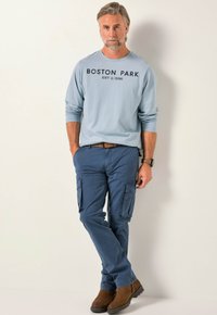 Svetle modrá košile s dlouhým rukávem, na které je v tmavých písmenech natištěno "BOSTON PARK", v kombinaci s modrými cargo kalhotami a hnědými kotníkovými botami.