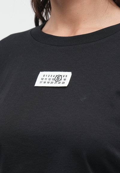 Fekete pamutpóló kerek nyakkal, elöl fehér szövetcímkével, rajta számokkal és egy kör alakú logóval.
