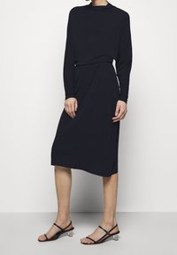 Femme vêtue d'une robe midi noire à manches longues avec un détail drapé, associée à des sandales noires à talons bas et à brides, debout sur un fond blanc.