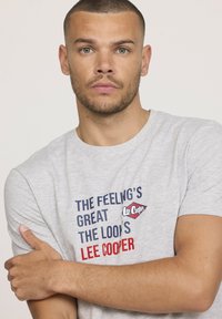 T-shirt en coton gris avec un texte imprimé bleu marine et rouge : "LA SENSATION EST GÉNIALE, LE LOOK EST LEE COOPER." Design à manches courtes et col rond.
