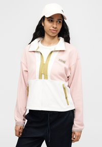 Νεαρή γυναίκα με ροζ και άσπρο fleece μπουφάν με φερμουάρ και λευκό καπέλο μπέιζμπολ, που στέκεται μπροστά σε απλό γκρι φόντο.