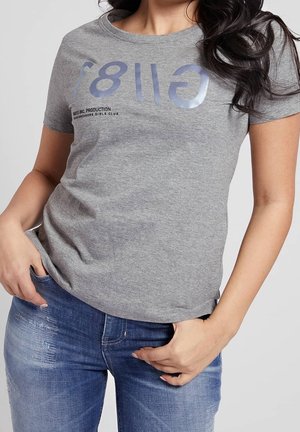 Kvinde iført en grå t-shirt med teksten "GIRLS" og blå falmede jeans, poserer med den ene hånd i lommen og den anden nær linningen.
