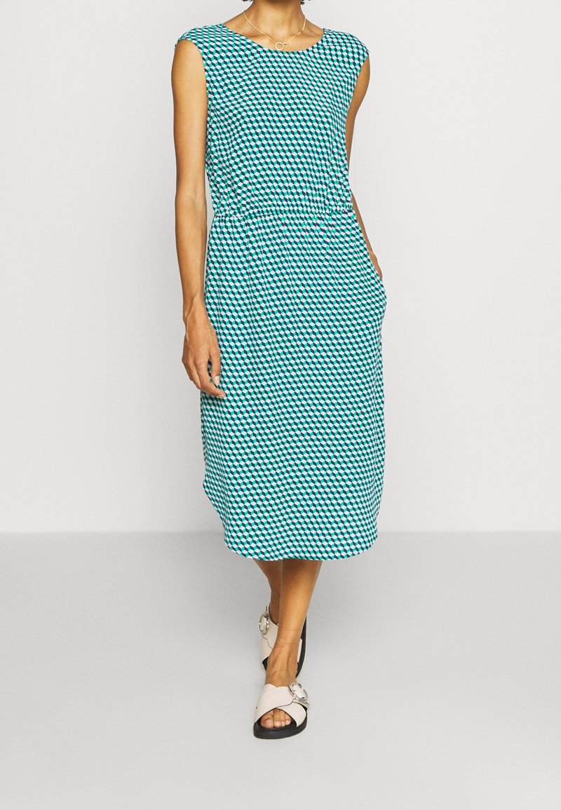Robe sans manches longueur au genou avec motif géométrique bleu, noir et blanc, taille élastique, poches latérales, portée avec des sandales rose clair à boucle argentée.