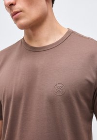 Νεαρός άνδρας που φοράει μια απλή καφέ μπλούζα με στρογγυλή λαιμόκοψη και ένα μικρό ανάγλυφο κυκλικό λογότυπο στο αριστερό μέρος του στήθους.