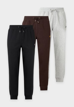 Homme debout portant un sweat à capuche zippé marron foncé et un pantalon de jogging assorti avec des baskets blanches sur un fond blanc uni.
