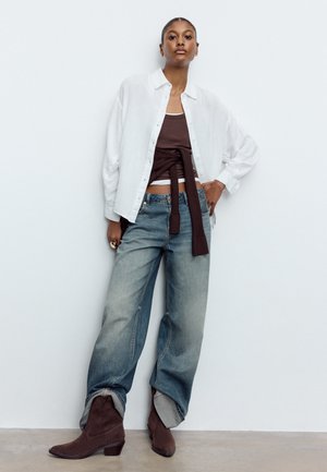 Frau trägt locker sitzendes weißes Hemd mit Knöpfen über braunem Oberteil mit gebundenen Ärmeln, weite Jeans mit hochgekrempelten Säumen und braune Stiefeletten, steht an einer Wand.