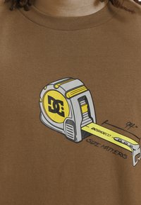Brown t-shirt featuring a graphic of a measuring tape in yellow and gray, with "DCSHOECO" and "SIZE MATTERS" text beneath.