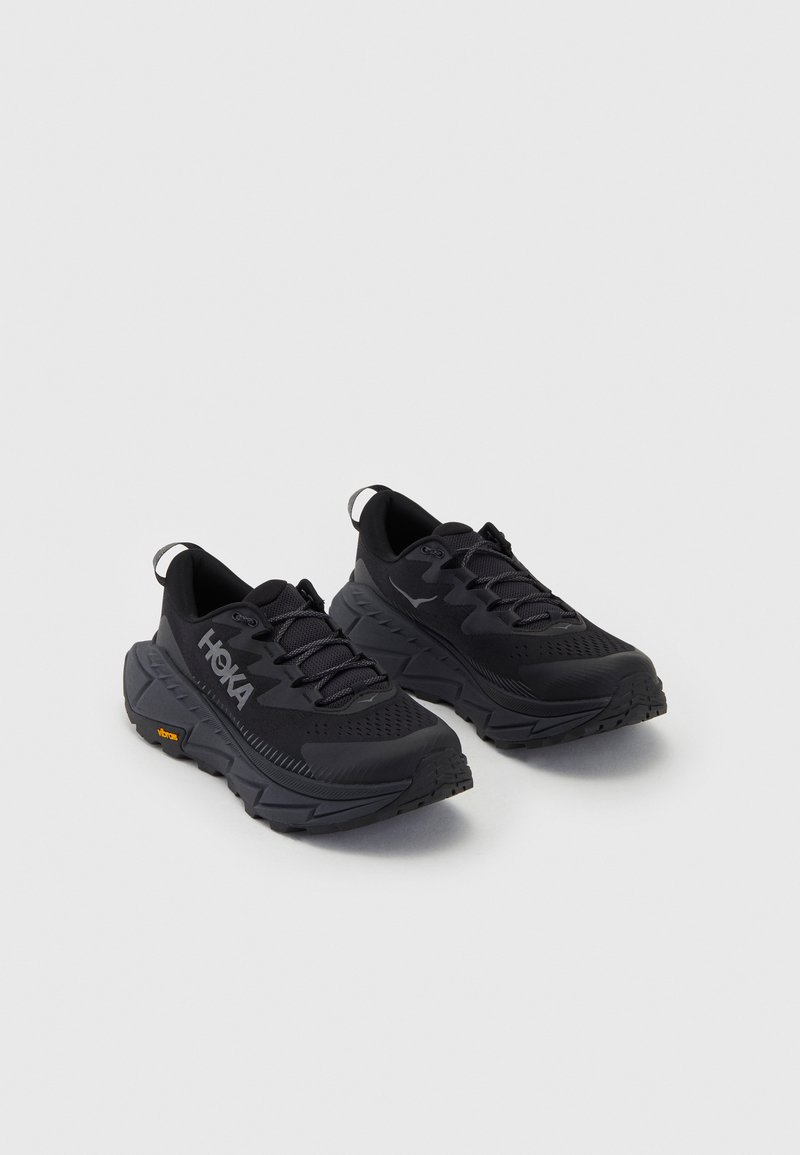 Sapatos de desporto pretos com um cabedal texturizado, almofada espessa e uma sola robusta. Logótipo HOKA visível, com realces amarelos vibrantes.