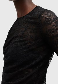 Gros plan d'une personne portant un haut moulant en dentelle noire avec des motifs floraux et un col rond, sur un fond clair uni.