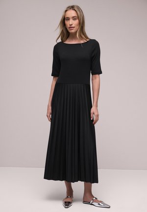 Žena s dlouhými blond vlasy, která má na sobě černé šaty s rukávy sahajícími po lokty a plisovanou sukní, stojící před jednobarevným světlým pozadím.