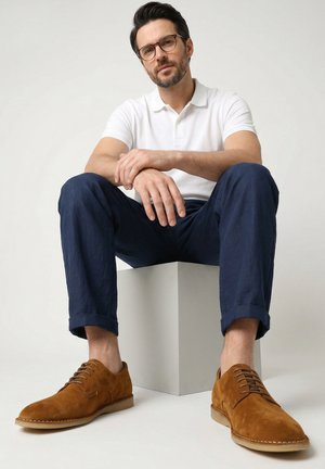 Man met bril zit op een witte kubus, draagt een wit poloshirt, donkerblauwe broek en bruine suède veterschoenen tegen een effen achtergrond.