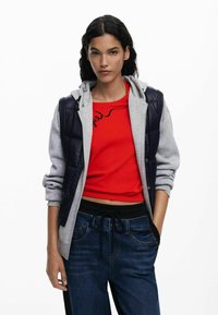 Grijze en marineblauwe puffer vest met een capuchon, laag over een rode cropped shirt. Gepaarde met donkere denim broeken met een trekkoord in de taille en zakken.